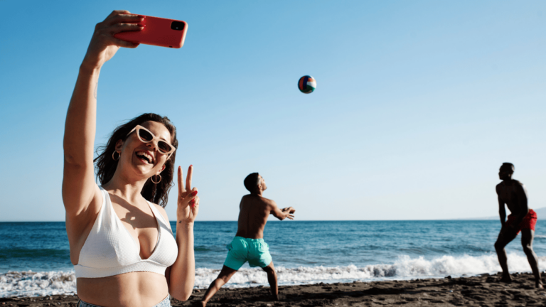 Woman taking a selfie while friends play volleyball during a trip.